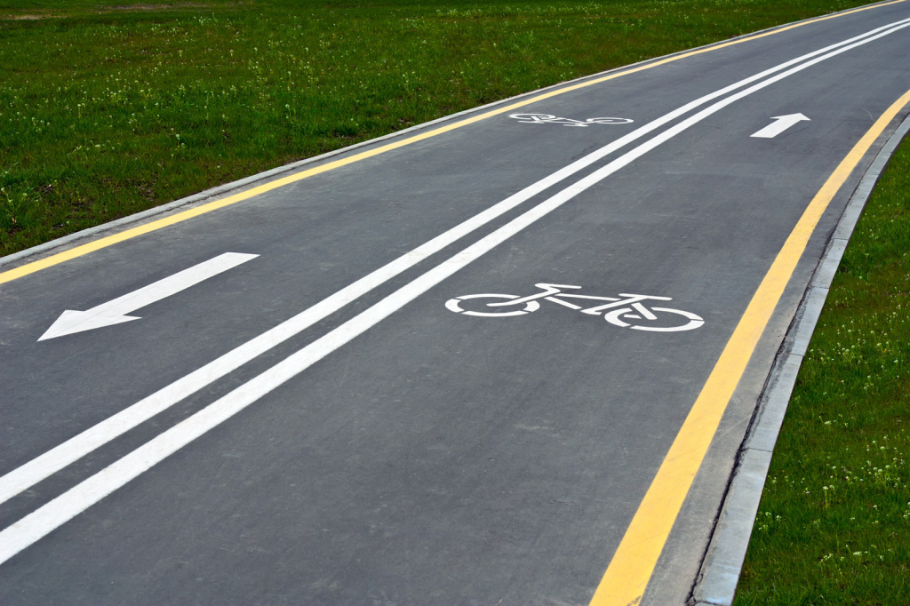 Une piste cyclable à double sens avec des lignes blanches au sol. Il y a des flèches indiquant la direction à suivre, ainsi que des symboles de vélo pour signaler que c'est une voie réservée aux cyclistes. De chaque côté de la piste, on peut voir de l'herbe.